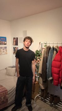 Νέος άντρας με μαύρη μπλούζα και σκούρο παντελόνι στέκεται σε ένα υπνοδωμάτιο με κρεβάτι, φωτογραφίες στον τοίχο, φυτό και μια κρεμάστρα με μπουφάν και παπούτσια.