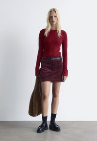 Rotes geripptes Langarmoberteil kombiniert mit einem bordeauxfarbenen Cordminirock, schwarzem Gürtel, dicken schwarzen Schuhen und knielangen schwarzen Socken. Fellmantel drapiert.