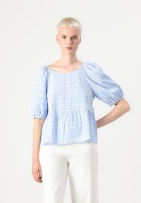 Lys blå og hvit rutete bluse med puffede ermer, rynket midje og rund hals, kombinert med hvite bukser.