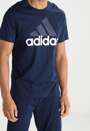 Mann trägt dunkelblaues Adidas-T-Shirt und passende Hose, steht vor einfachem hellem Hintergrund.