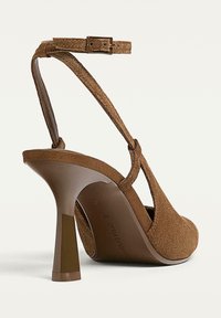 Scarpa con tacco alto in suede marrone, con cinghia alla caviglia e punta arrotondata, mostrata da dietro e di lato su uno sfondo bianco.