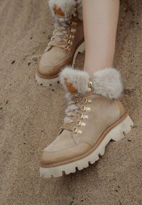 Botas de tobillo de ante beige con borde de pelo, herrajes dorados y una suela gruesa y estriada. Los cordones y una etiqueta decorativa completan el diseño.