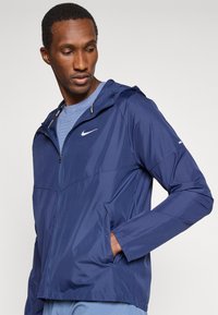 Navy lightweight jacket with a zip closure, hood, side pockets, and Nike logo on the chest. Smooth texture and athletic fit.