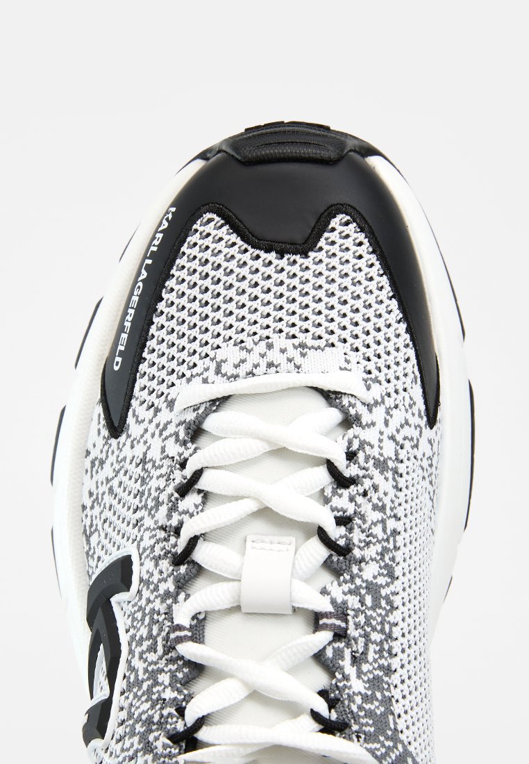 Primer plano de una zapatilla tejida negra, blanca y gris con cordones blancos y puntera de goma, con la marca "Karl Lagerfeld" en el panel lateral.