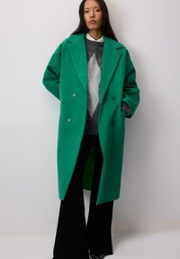 Roheline villaseguse topeltnööbiga mantel, millel on ülepaisutatud kraed, kaks eesmistTaskut ja mugav lõige, koos halli mustriga sviitriga ja mustade pükstega.