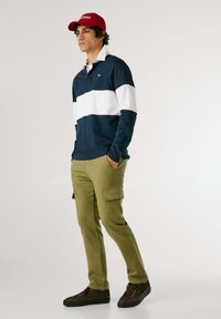 Marinblå og hvide stribede langærmede polo shirt, parret med olivengrønne cargo bukser. Outfitet har en rød cap og brune sneakers med sorte såler.