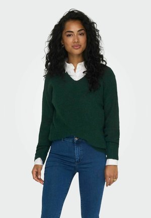 Mörkgrön, stickad tröja med V-hals och avslappnad passform, över en vit skjorta med krage, kombinerad med blå jeans. Texturen är mjuk.
