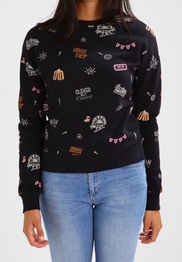 Femme portant un sweat-shirt noir avec un texte coloré et de petites icônes graphiques, assorti à un jean bleu clair.