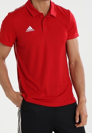 Homme portant un polo rouge à manches courtes Adidas avec un pantalon noir à rayures blanches, debout avec une main dans la poche.