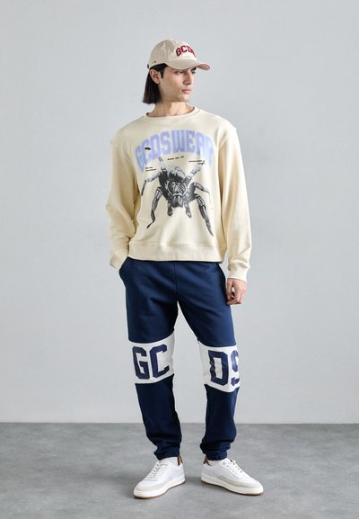 Sweatshirt crème avec un imprimé graphique d'araignée, associé à un jogging bleu marine avec des inscriptions blanches en gras "GC" et "09", complété par des baskets blanches.