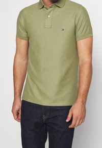 Homme portant un polo à manches courtes olive avec un petit logo sur la poitrine et un jean en denim bleu foncé, se tenant devant un fond uni.