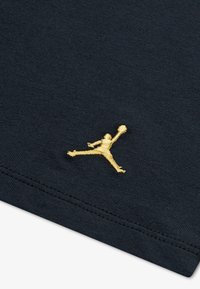 Tissu noir avec un logo Jumpman brodé en or. Le matériau est lisse, avec une texture fine et des coutures nettes le long des bords.