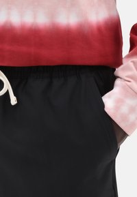 Sudadera de teñido anudado roja y rosa con cordón; combinada con pantalones cortos negros con bolsillos laterales y una textura suave.
