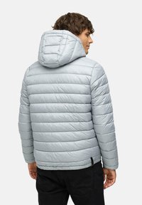 Lyseblå, polstret jakke med hætte, med horisontal quiltning, en blank overflade og en sort justerbar detalje forneden.