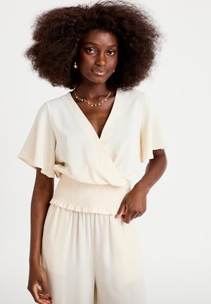 Kobieta o dużych kręconych włosach, ubrana w kremową bluzkę wiązaną na kopertę i pasujące spodnie, dopełniona delikatnym naszyjnikiem i kolczykami.