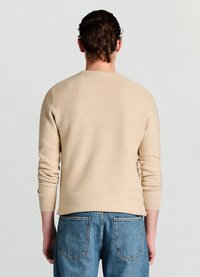 Beiger Rippstrickpullover mit langen Ärmeln und Rundhalsausschnitt, kombiniert mit blauen Jeans mit Gesäßtaschen.