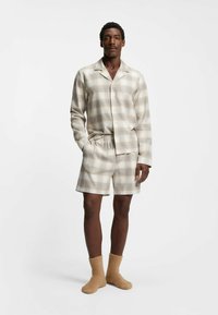 Beigeaktige, leichte karierte Schlafanzug-Set, bestehend aus einem knopfbaren Hemd und Shorts, aus weichem Stoff. Kombiniert mit tanfarbenen, flauschigen Socken.