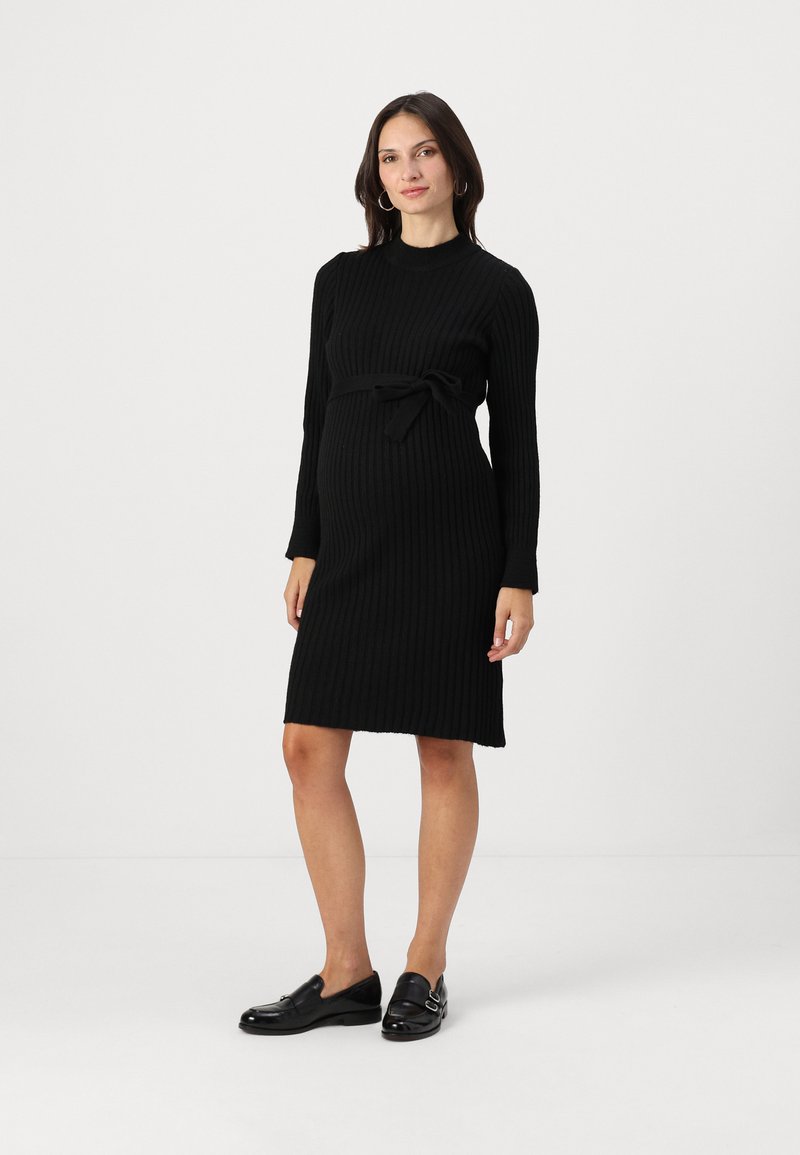Svart ribbestrikket kjole med lange ermer, sløyfebelte ved midjen, og knelangs lengde. Kombinert med svarte slip-on sko.