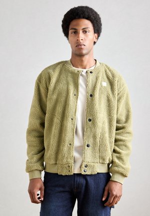 Giacca in fleece verde oliva chiaro con superficie testurizzata, polsini a coste e chiusura con bottoni a pressione, indossata sopra una camicia bianca e jeans.