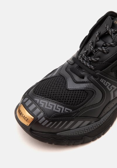 Μαύρο sneaker Versace με λεπτομέρειες από δίχτυ και δέρμα, διακοσμητικά σχέδια, αντανακλαστικές ρίγες και χρυσή μεταλλική πινακίδα με το λογότυπο στο δάχτυλο.