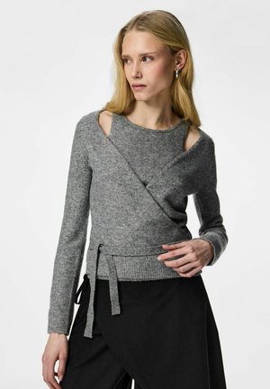 Mujer con cabello rubio largo que lleva un suéter gris tejido con aberturas en los hombros y una falda negra asimétrica, posando contra un fondo blanco.