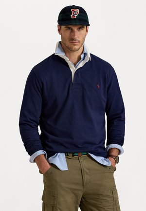 Marineblauwe poloshirt met lange mouwen en een witte kraag. Groen-zwart geblokt petje. Tan cargo broek met zakken. Bruine leren riem, zilveren horloge.