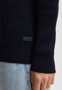 Maglione lavorato a maglia in navy con texture a coste, orlo aderente e etichetta nera con "STRELLSON". Il design del maglione è affusolato, aggiungendo dettagli.