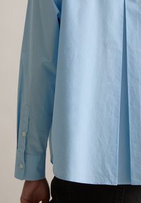 Camicia azzurro chiaro con vestibilità comoda, polsini con bottoni e piega posteriore. Il tessuto appare liscio e leggermente stropicciato.