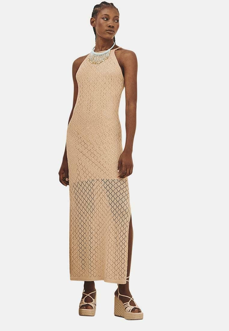 Vestido largo de punto en beige con un patrón de diamantes, escote halter, aberturas laterales y complementado con un collar de múltiples hilos. Lleva unas cuñas de plataforma.