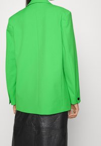 Blazer verde con spalle strutturate, una fessura posteriore singola e bottoni neri sui polsini, indossato su una gonna di pelle nera.