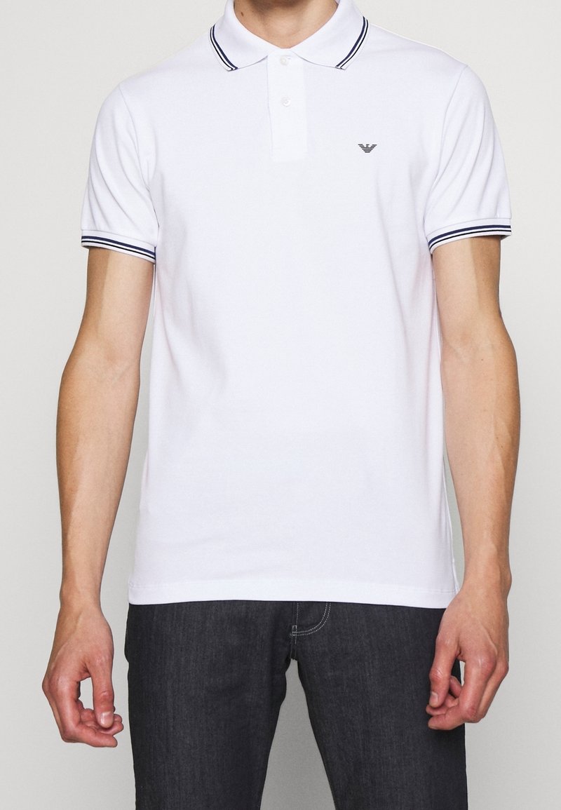 Homme portant un polo blanc à manches courtes avec des rayures marines sur le col et les manches, associé à un jean foncé.