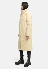 Beige langer Puffercoat mit hochgeschlossenem Kragen, seitlichen Taschen und bestickten Details am Ärmel. Trägt er über schwarzen Jeans und Kampfstiefeln.