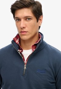 Felpa blu navy con zip e colletto alto, caratterizzata da un logo sottile e una camicia di flanella a quadri rossi indossata sotto. Texture del tessuto liscia.