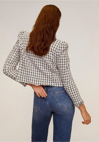 Giacca cropped a scacchi bianca e nera con maniche lunghe, caratterizzata da un tessuto testurizzato e dettagli sulle spalle, abbinata a jeans in denim blu.
