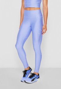 Žena nosící světle fialové legíny s vysokým pasem a bočním kapsou, v kombinaci s odpovídajícím sportovním podprsenkovým topem a modro-černými běžeckými botami Puma.
