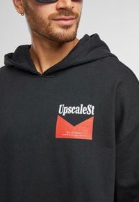 Felpa con cappuccio nera caratterizzata da un prominente logo grafico rosso e bianco con il testo "UpscaleSt". Tessuto morbido con vestibilità comoda.