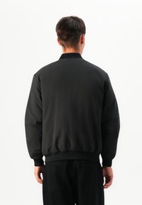 Mann in einer schwarzen Bomberjacke und schwarzen Hosen, der mit dem Rücken zu einem schlichten weißen Hintergrund steht.