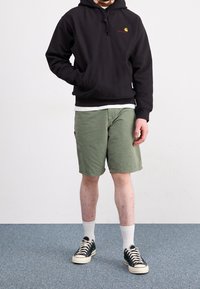 Schwarzer Kapuzenpullover mit vorderer Tasche, olivgrüne Shorts, weißes T-Shirt darunter, weiße Socken und schwarze Sneakers mit weißer Sohle.