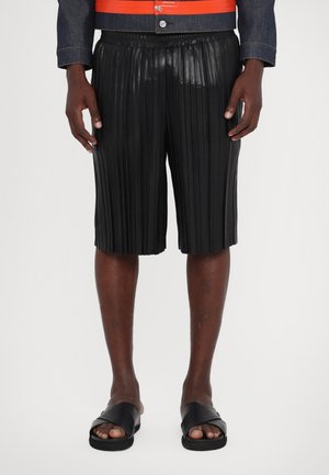 Schwarze plissierte Shorts aus glänzendem Material, mit einem elastischen Bund und einem knielangen Schnitt. Schwarze Schlupfsandalen runden den Look ab.
