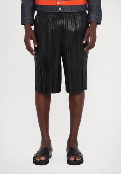 Black pleated shorts made of a shiny material, featuring an elastic waistband and a knee-length cut. Black slip-on sandals complete the look.
