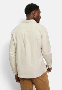 Light beige long-sleeve shirt with a back pleat, button cuffs, and a classic collar, worn with brown corduroy pants. Smooth fabric texture.