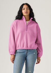Pull polaire rose avec col à demi-zip, manches longues, détails aux poignets côtelés et coupe décontractée. Associé à un jean bleu clair déchiré.