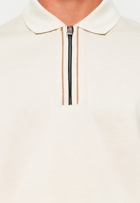 Poloshirt i off-white stoff med kort glidelås og krage. Har en sentral vertikal stripe med svart og brun detaljer.