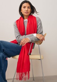 Écharpe rouge aux bords frangés drapée sur une personne assise, portée avec un pull gris et un jean bleu, sur un fond neutre.