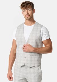Chaleco gris claro con un sutil patrón de cuadros, dos bolsillos frontales y un cierre de tres botones, llevado sobre una camiseta blanca.