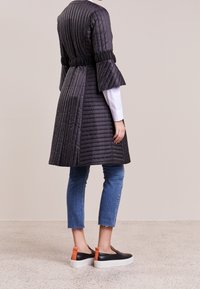 Svart quiltad kappa med skärp i midjan och vida ärmar, kombinerad med korta blå jeans och svarta slip-on-skor med orange detaljer.