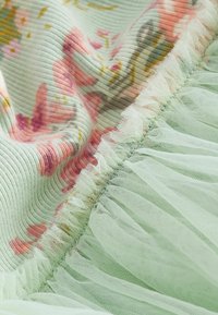 Látka s květinovým vzorem v jemné mátové zelené barvě, s jemně strukturovaným povrchem a decentními tylovými pruhy podél okraje pro vrstvený detail.