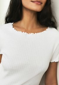 Weißes, geripptes Top mit einem gezackten Ausschnitt und kurzen Ärmeln mit Rüschensaum, gefertigt aus einem weichen, elastischen Stoff.