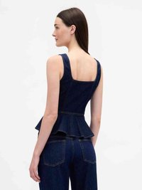 Donna con capelli castani lisci che indossa un top in denim blu scuro senza maniche con orlo peplum e pantaloni in denim a vita alta coordinati, di spalle.