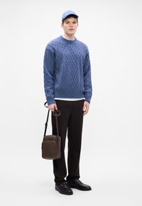 Lichtblauwe kabelgebreide trui, donkere broek, zwarte schoenen en blauwe pet. Bruine schoudertas met een gladde textuur en rechthoekige vorm.